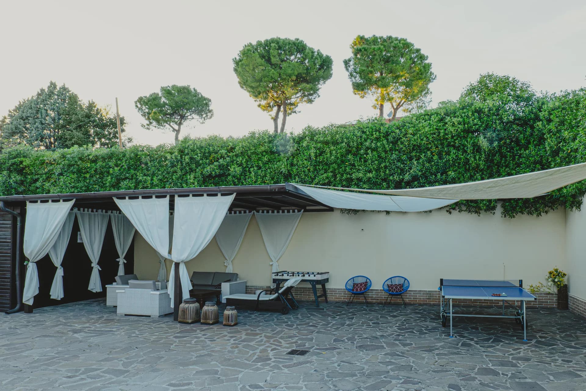 Area relax esterna con gazebo dalle tende bianche, divani, lettini, calcio balilla e tavolo da ping-pong sul cortile in pietra
