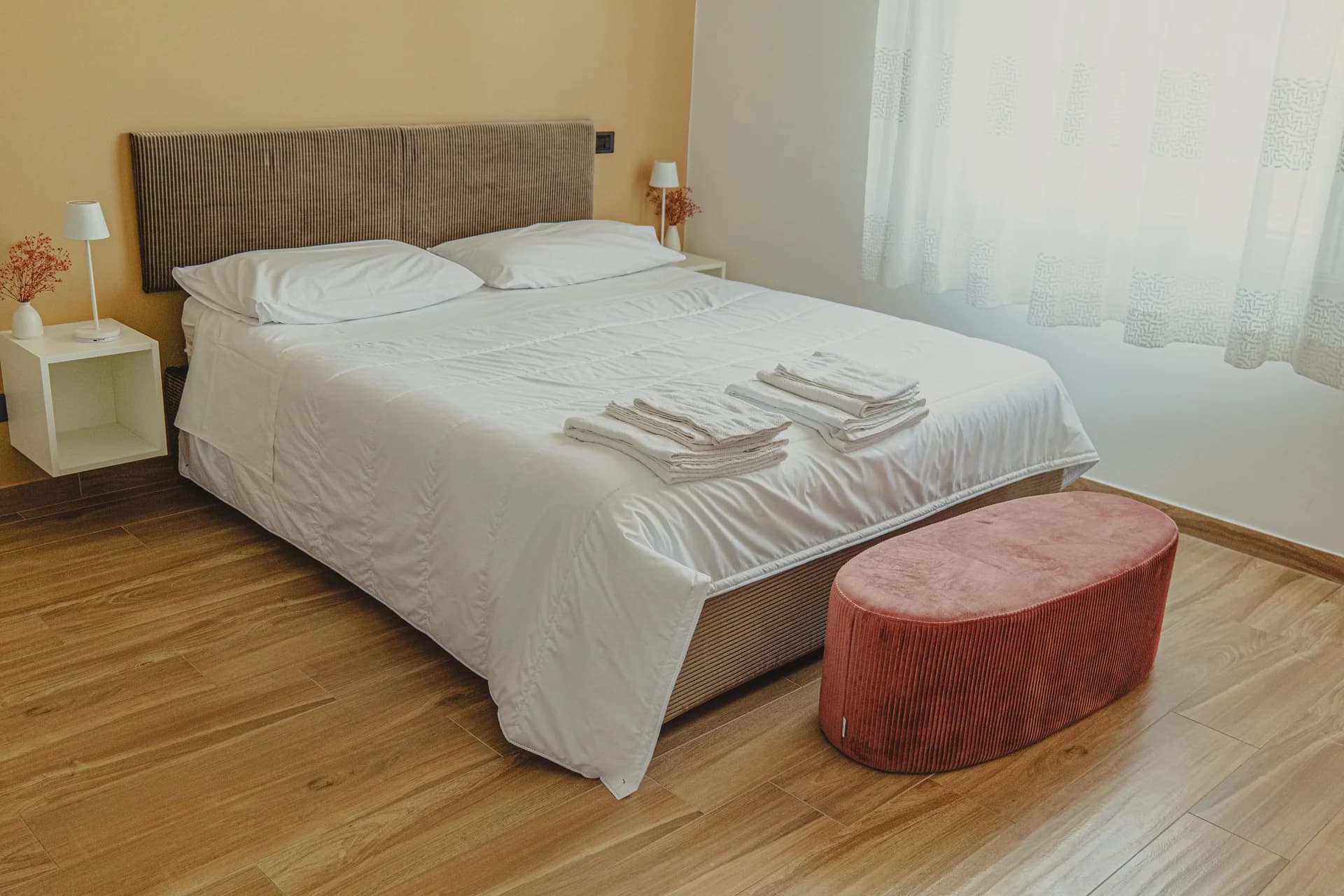 Camera matrimoniale luminosa con letto bianco, testata a coste, parquet, pouf in velluto terracotta e tende chiare