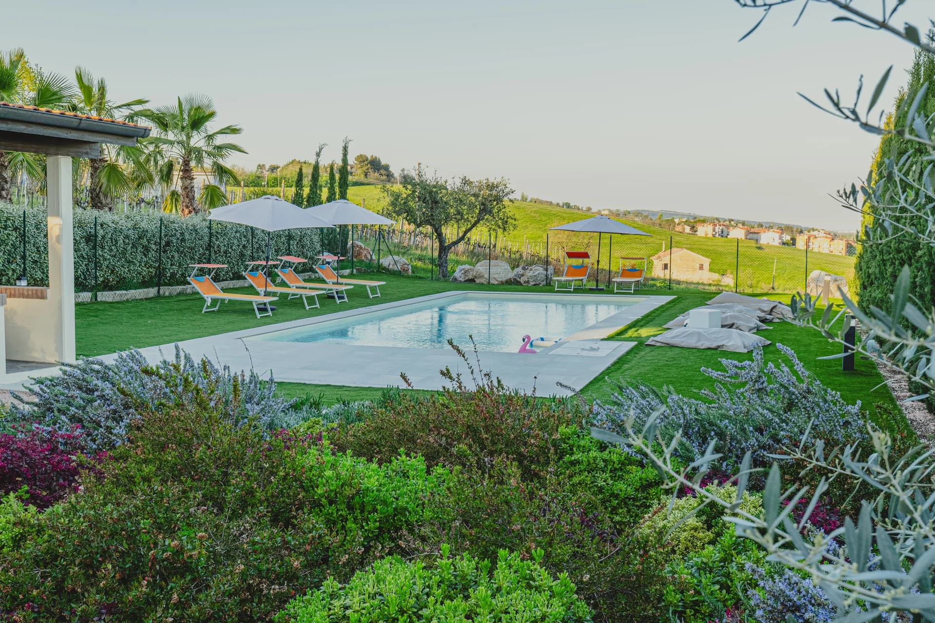 La piscina affacciata sul panorama delle colline, con lettini, ombrelloni e giardino fiorito in primo piano