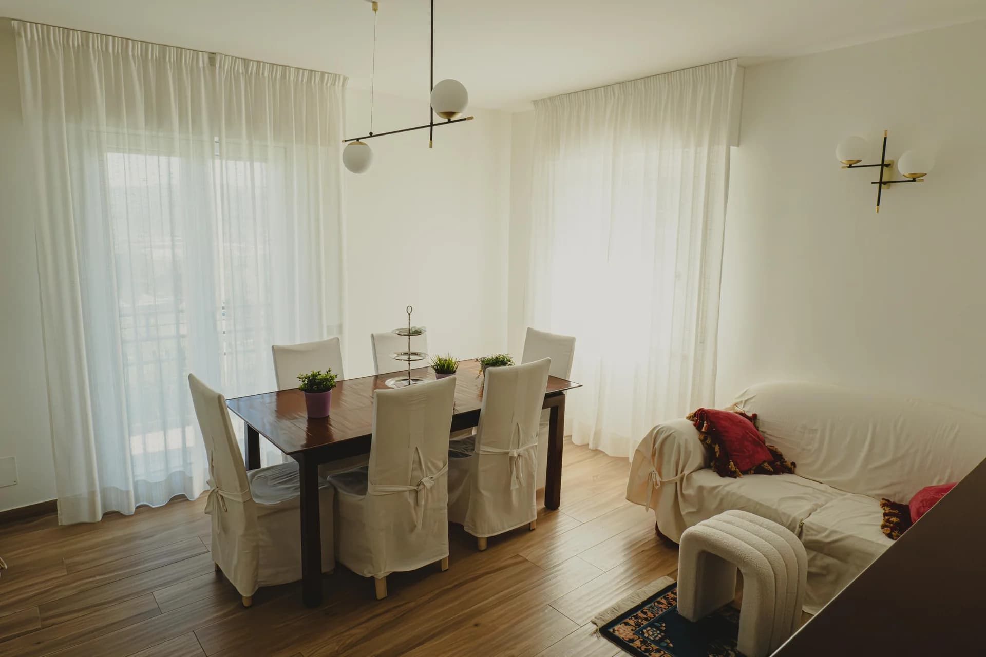 Sala da pranzo con tavolo in legno e sedie vestite di bianco, lampadario a sfere e divano con cuscini nel soggiorno luminoso