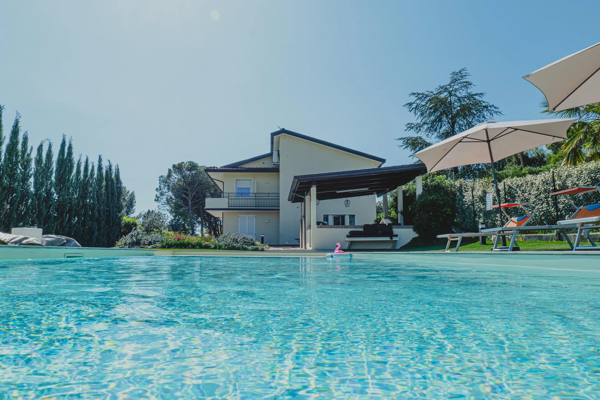 La villa vista dal filo dell'acqua della piscina, tra cipressi e ombrelloni, in una giornata di sole